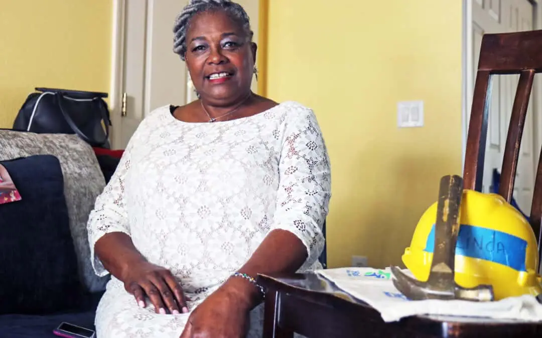 A woman sits on a couch and smiles at the camera, a hard hat and hammer next to her